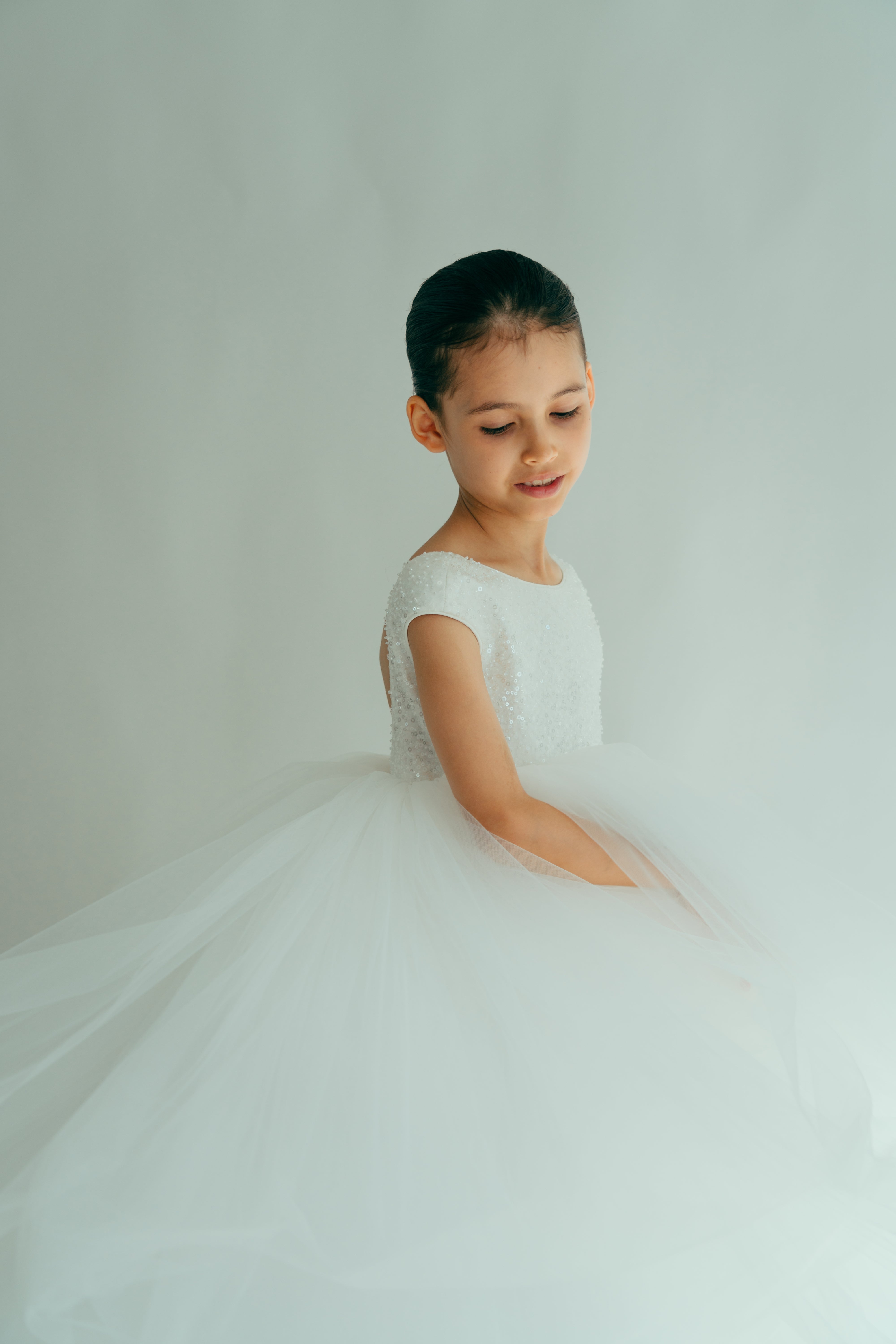A young girl in a white, maxi dress adorned with sequins and beads, posing in a semi-profile stance.