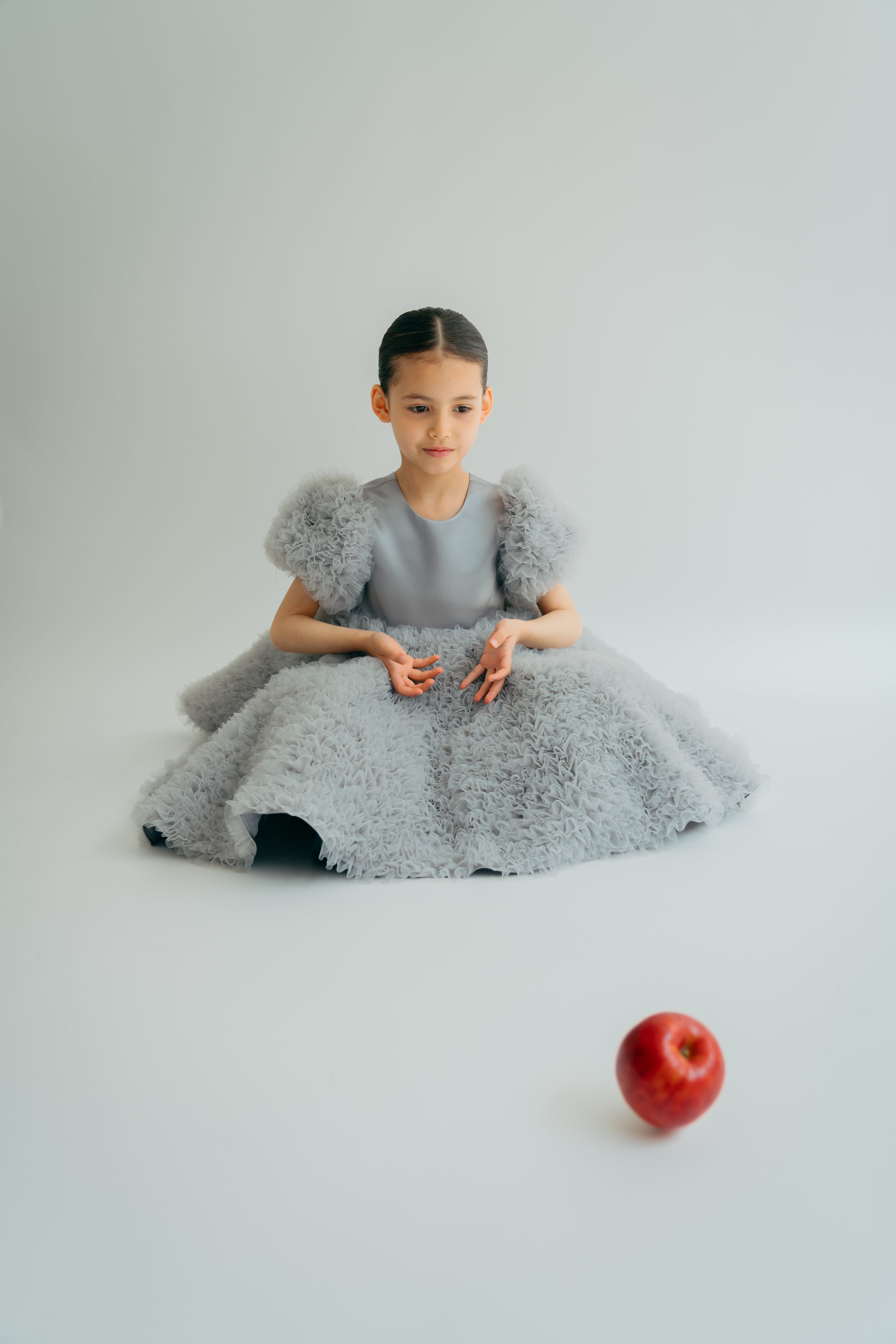 A young child wearing a grey dress with a voluminous skirt and puff sleeves, sitting next to a red apple.