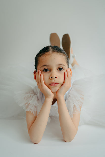 A young child wearing a white tulle dress with pintucks, posing with hands on cheeks.